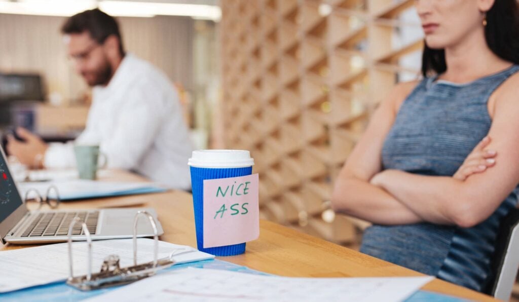 Woman sitting at her work desk with a post-it note placed on her coffee cup that reads "Nice ass"