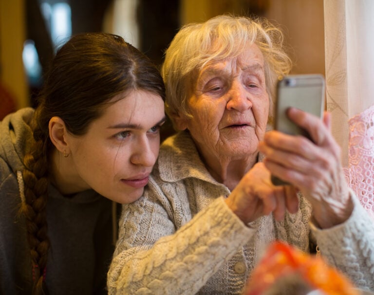 An elderly woman looks at a smartphone, with her adult granddaughter.