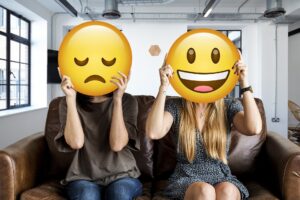 Two people sitting on a couch, one holding a mask with a sad face emoji, the other holding a mask with the happy face emoji